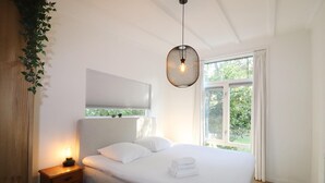 Een slaapkamer met een bed, nachtkastje, hanglamp, raam en uitzicht op groen buiten.