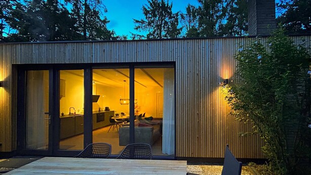 Een modern huis met grote schuifdeuren die leiden naar een goed verlicht interieur, een terras met zitplaatsen en een houten gevel.