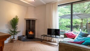 Een gezellige woonkamer met een open haard, een televisie op een dressoir, een bank met kleurrijke kussens, en een groot raam met een parasol.