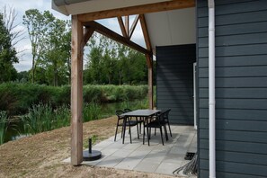An outdoor patio with a table and chairs, wooden beams, and a view of a pond and trees. - Nice holiday home near the waterfront (Zuid-Holland)