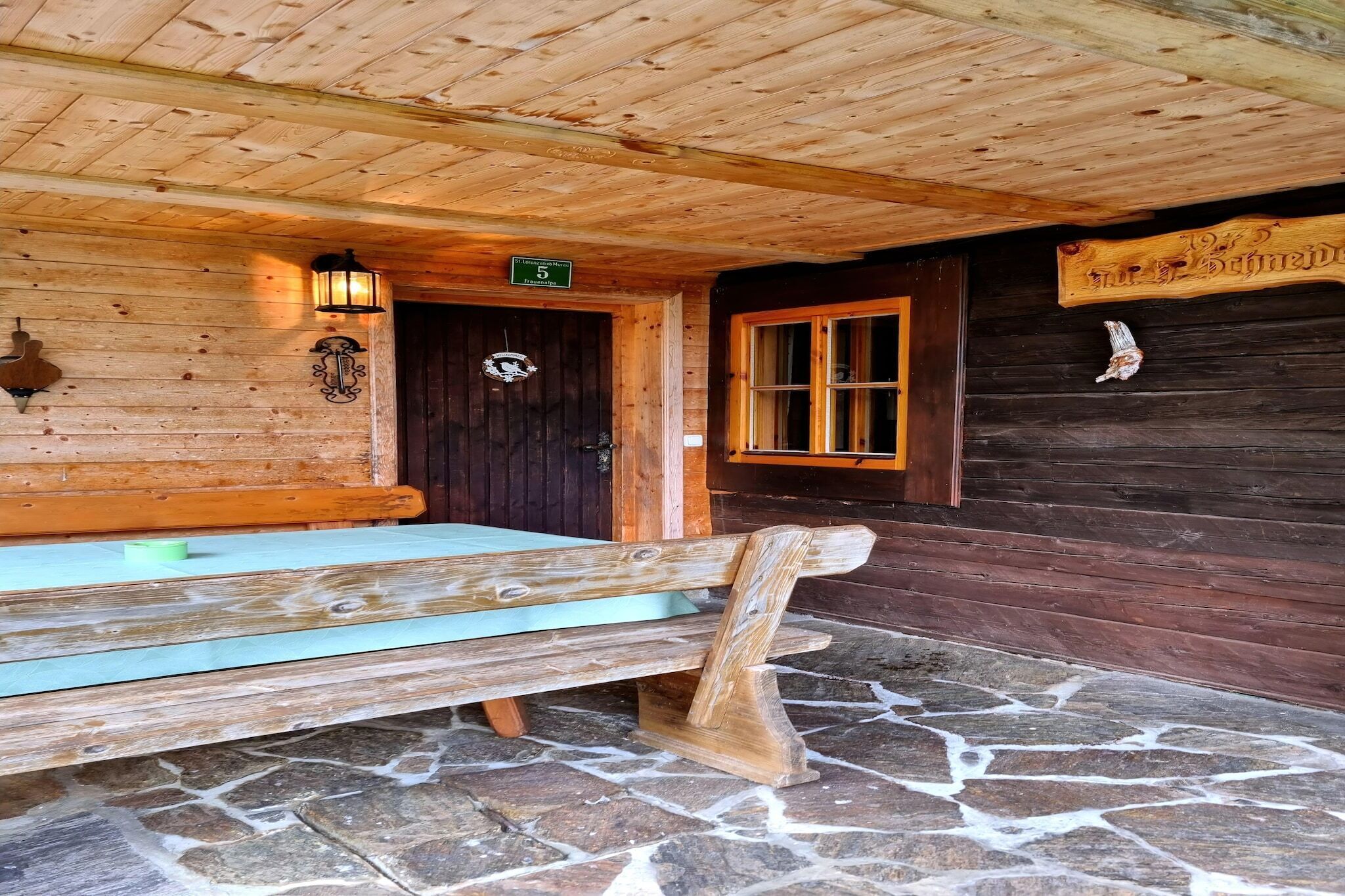 Una cabaña de madera con piso de piedra, un banco en el exterior y una puerta con un cartel encima.
