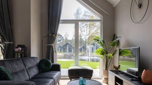Un salon moderne comprenant un canapé, un fauteuil, une table basse et un téléviseur posé sur un meuble en bois. Une grande fenêtre offre une vue sur un jardin et des constructions extérieures.