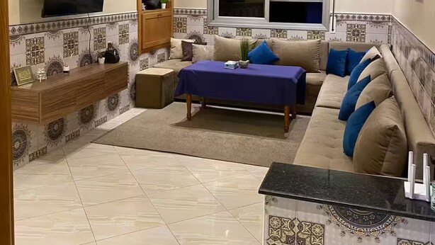 A living room with a sofa, a TV mounted on the wall, a window with shutters, and a decorative ceiling.