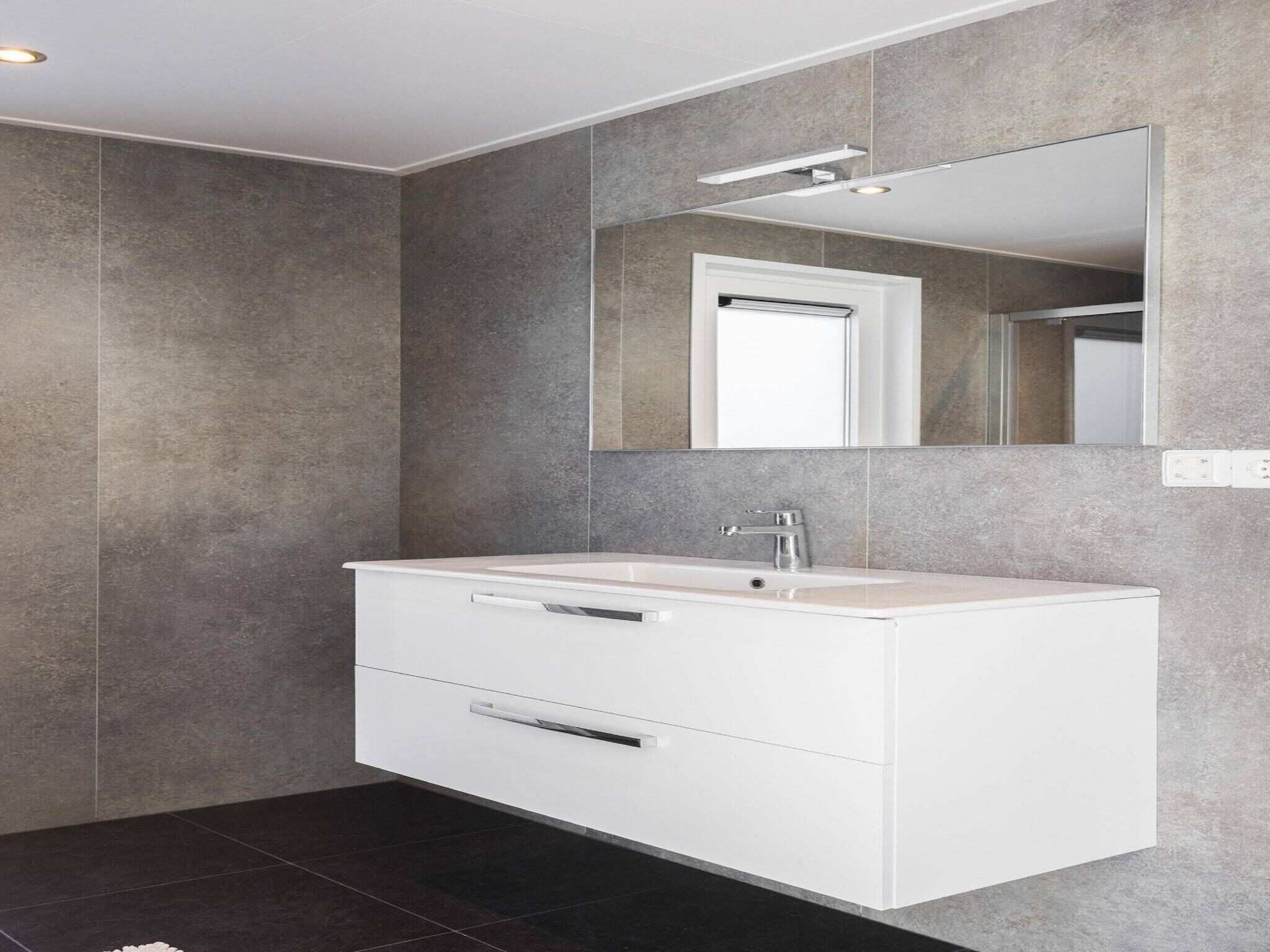 Une salle de bain moderne avec un meuble-lavabo blanc, un grand miroir et des murs recouverts de carreaux gris.