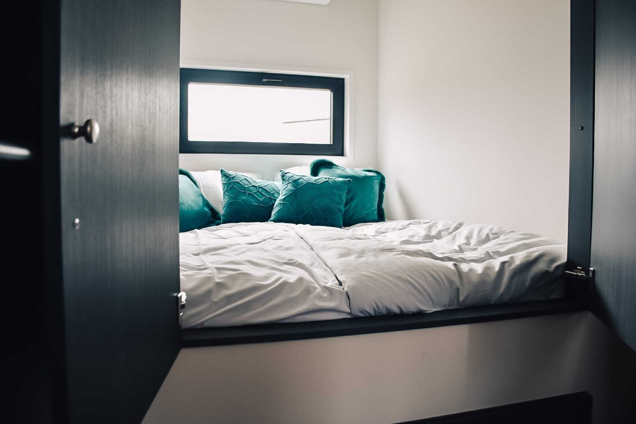 A bed with white bedding and teal pillows, a window above it, and a door on the left.