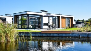 Une maison moderne dotée de grandes fenêtres en verre, d’un patio avec des meubles d’extérieur et d’un petit étang au premier plan.