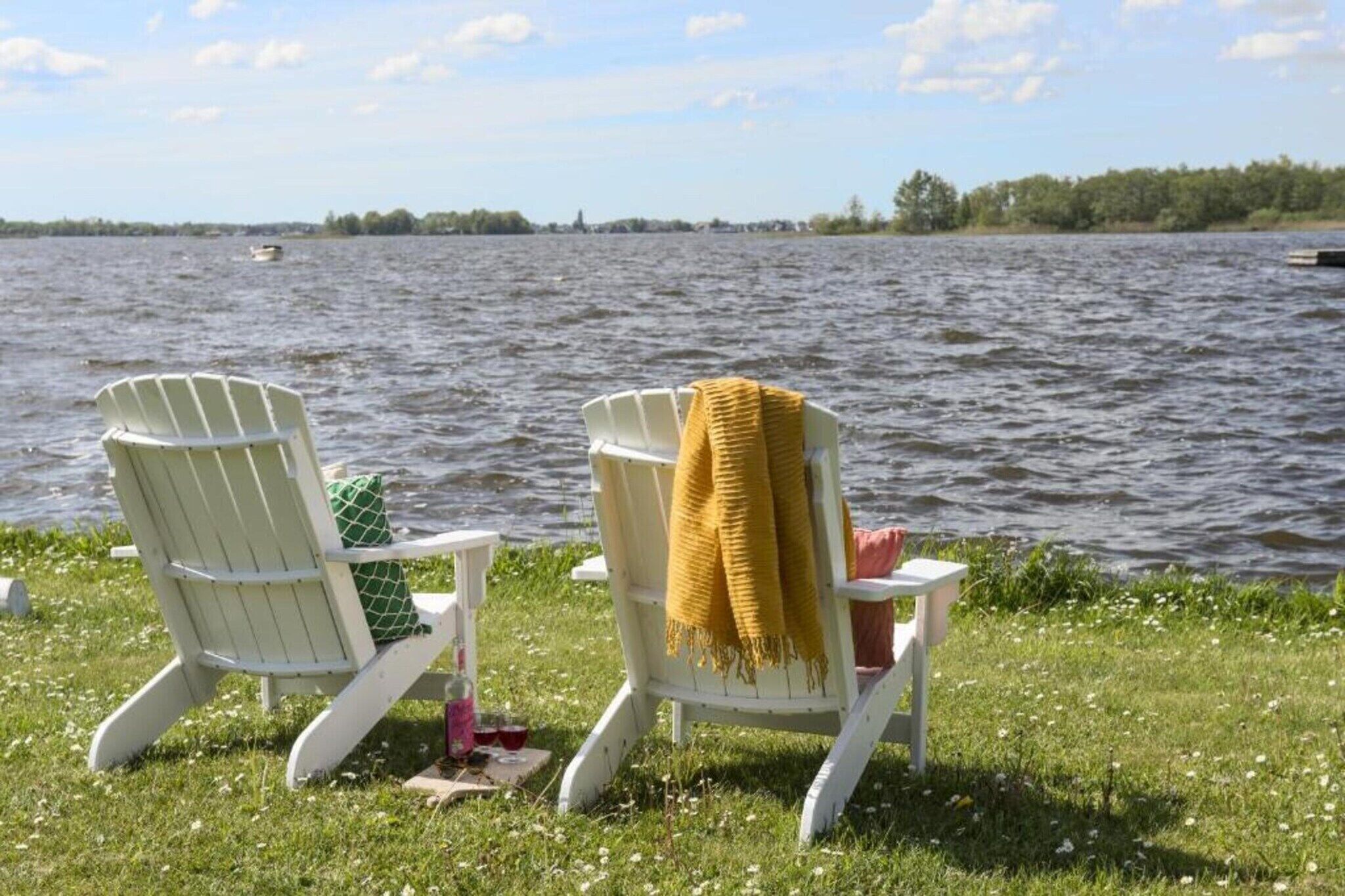 Deux fauteuils Adirondack placés près d’un lac, avec une zone herbeuse et un bateau visible au loin.