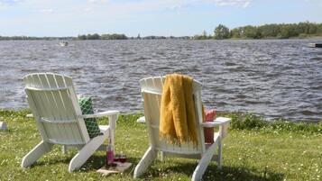 Deux fauteuils Adirondack placés près d’un lac, avec une zone herbeuse et un bateau visible au loin.