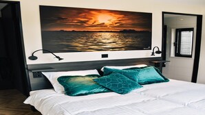 Une chambre avec un grand lit, deux coussins de couleur turquoise, une table de chevet, une lampe fixée au mur et un tableau représentant un coucher de soleil au-dessus du lit.