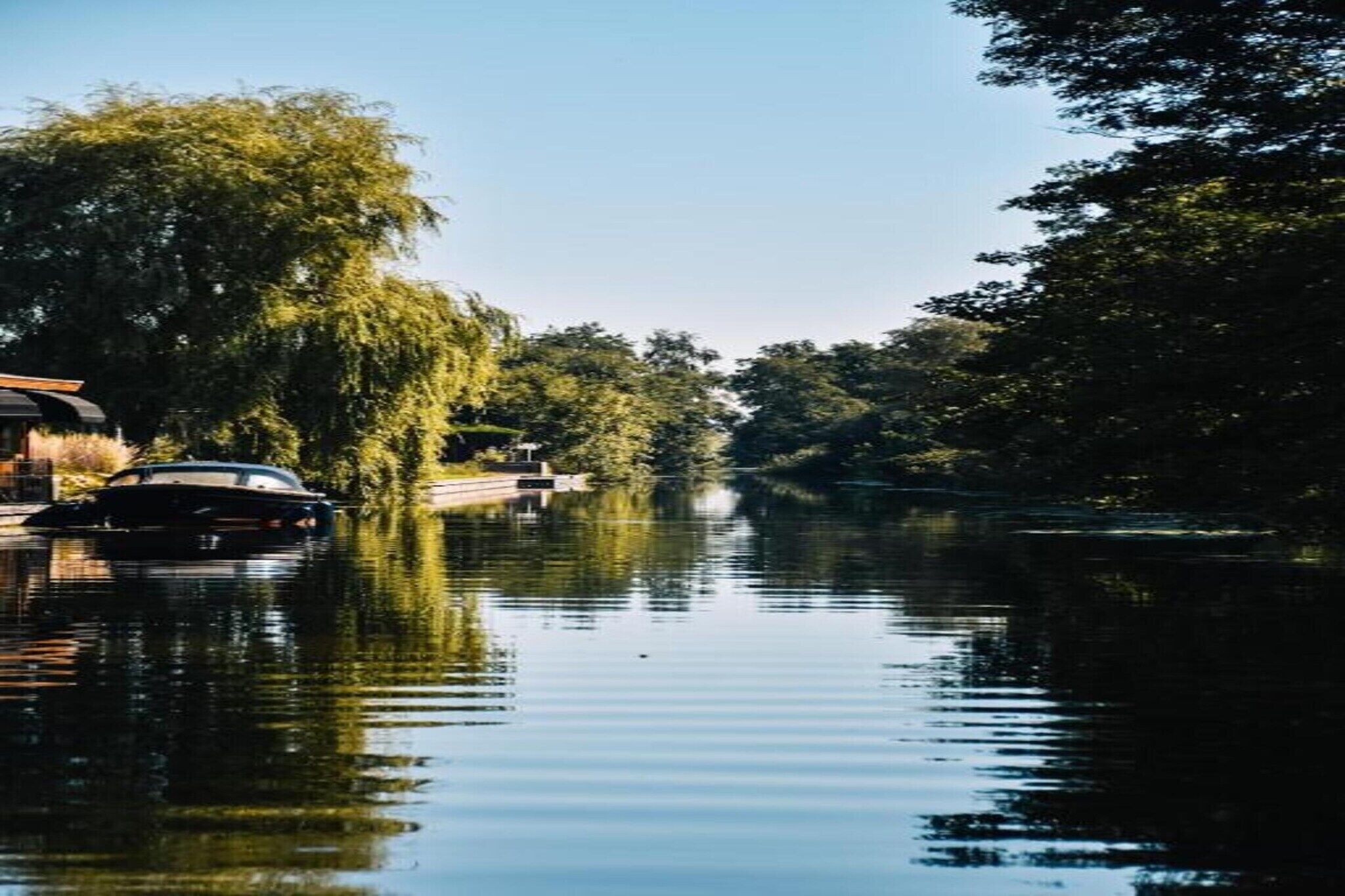 Un fiume tranquillo con una barca ormeggiata a sinistra, immerso nel verde e sotto un cielo sereno.