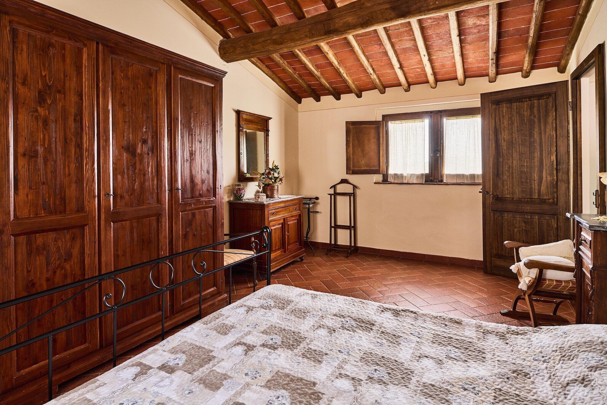 A bedroom with a large wooden wardrobe, a bed with a patterned bedspread, a wooden dresser, and a window with curtains.