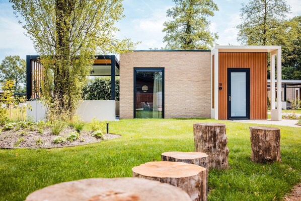 A modern building with a brick facade, a glass door, and a wooden door. There are trees and a grassy area with wooden stumps in the foreground. - Chalet in Veluwe near Hoge Veluwe Park (Voorthuizen)