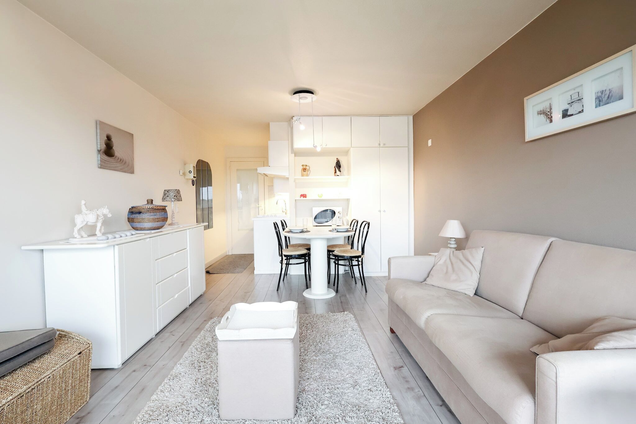 A modern living room with a sofa, a dining table with chairs, a kitchen area with cabinets, and a small decorative statue on the counter.