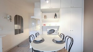 Un comedor con una mesa redonda, sillas, un armario blanco, un microondas y un estante pequeño con adornos.