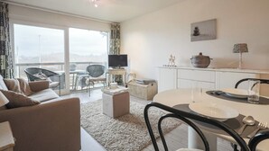 Una sala moderna con sofá, una mesa de comedor para dos, televisión y un balcón con vistas.