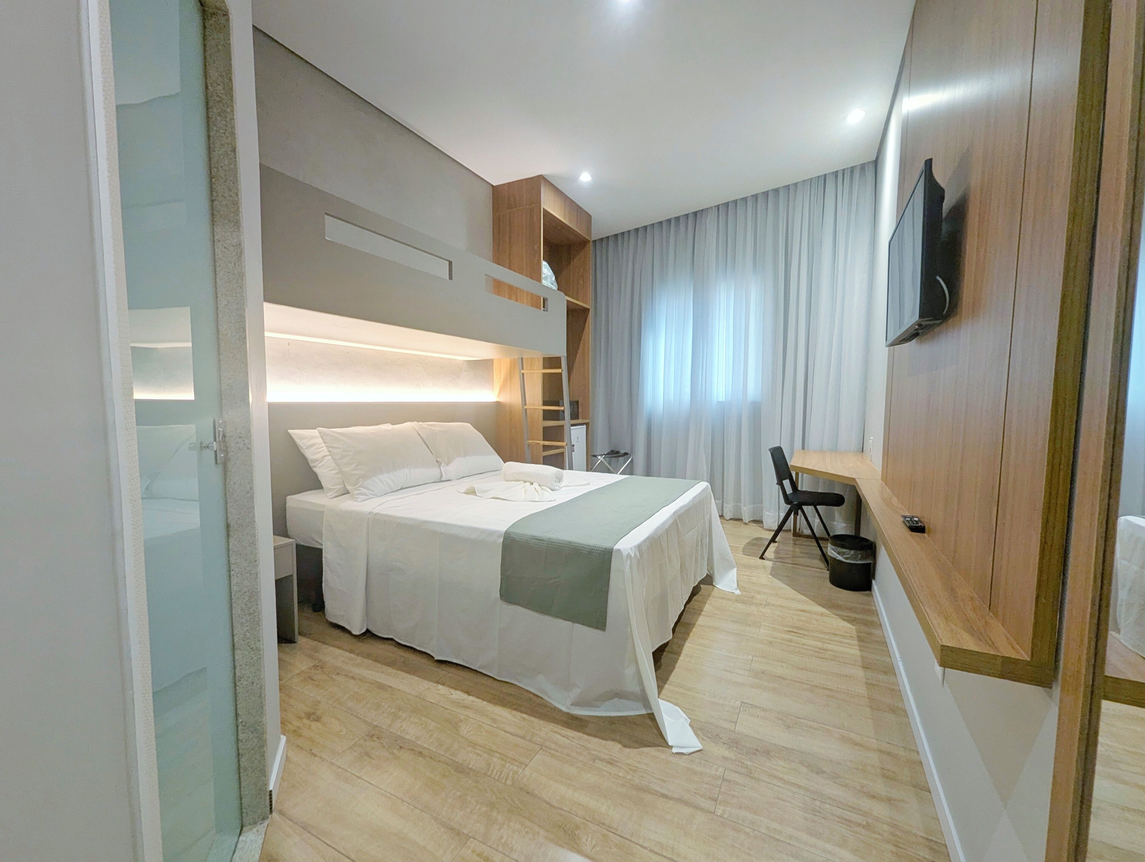 Quarto de hotel moderno com cama, escrivaninha, cadeira, televisão e janela com cortinas.
