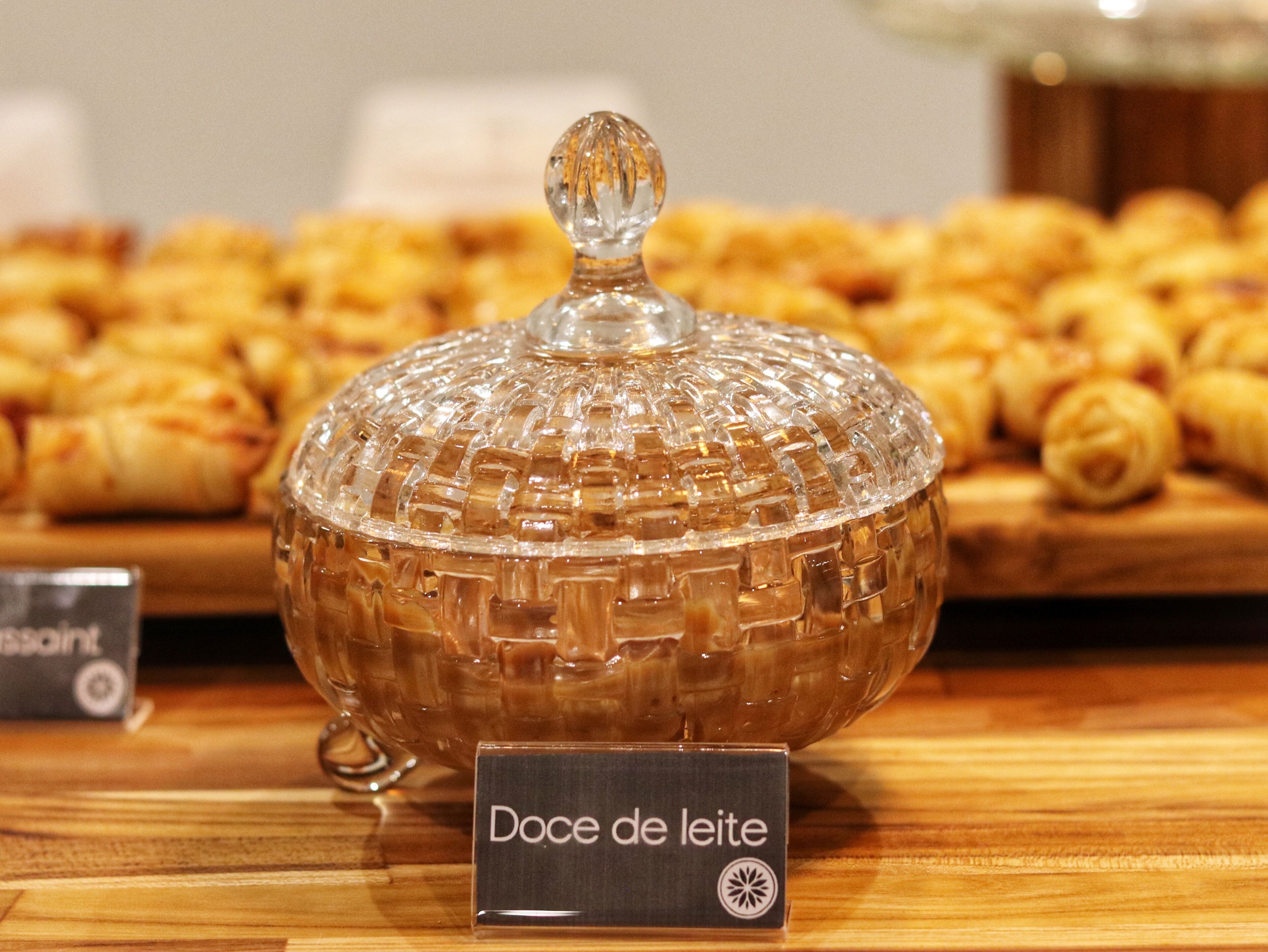 En glasburk märkt med texten ”Doce de leite” står på en träyta, med bakverk i bakgrunden.