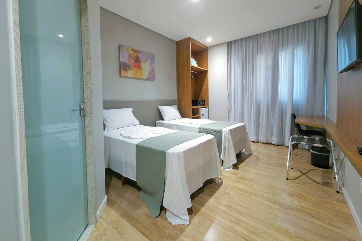 A hotel room with two beds, a desk, a chair, a TV, a wardrobe, and a painting on the wall.