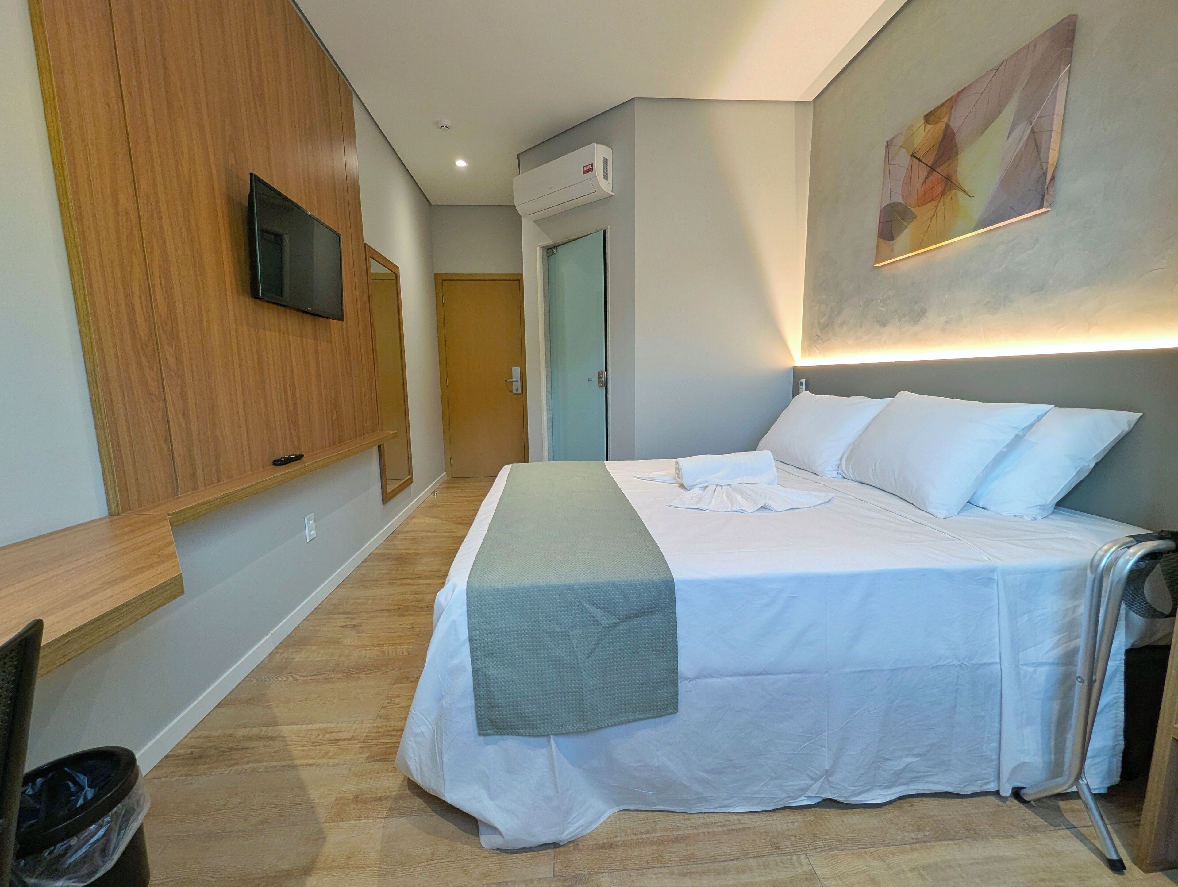 A hotel room with a bed, a TV mounted on the wall, a wooden panel, a door, and a painting on the wall.