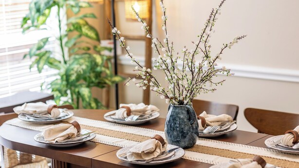 Une table à manger dressée avec des assiettes, des couverts et des serviettes, et un vase rempli de branches et de petites fleurs placé au centre.