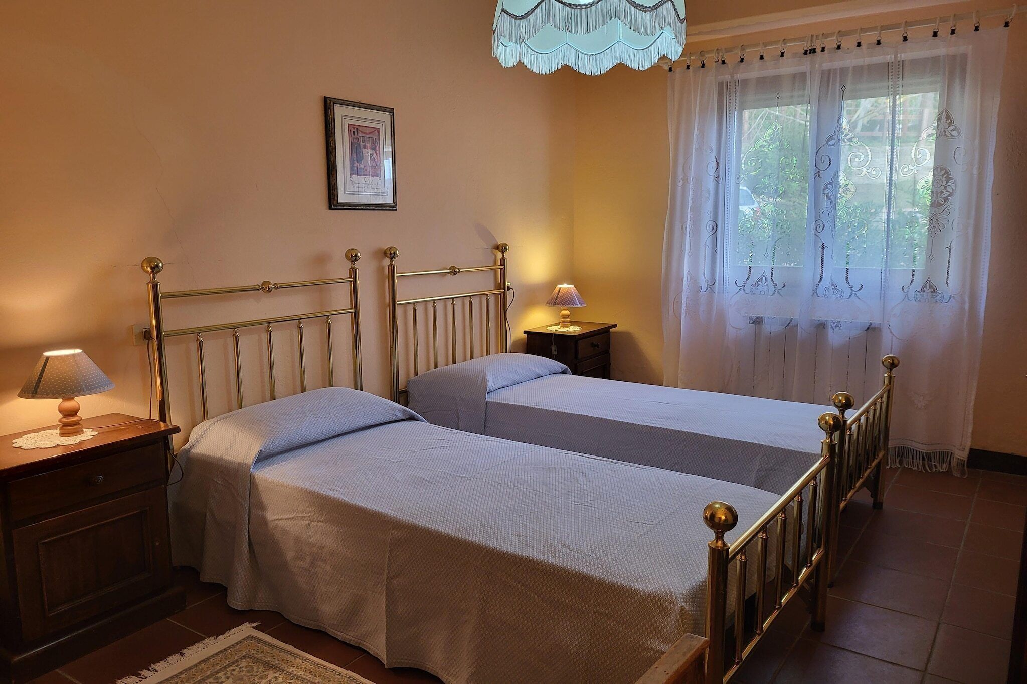 Ein Schlafzimmer mit zwei Einzelbetten, einem Nachttisch mit Lampe, einem gerahmten Bild an der Wand und einem Fenster mit durchsichtigen Vorhängen.
