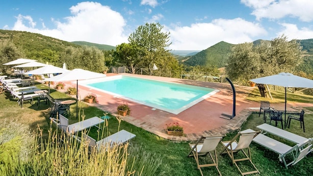 Una piscina rectangular con un patio circundante, equipada con sillones de descanso y sombrillas, frente a un telón de fondo de colinas verdes y un cielo despejado.