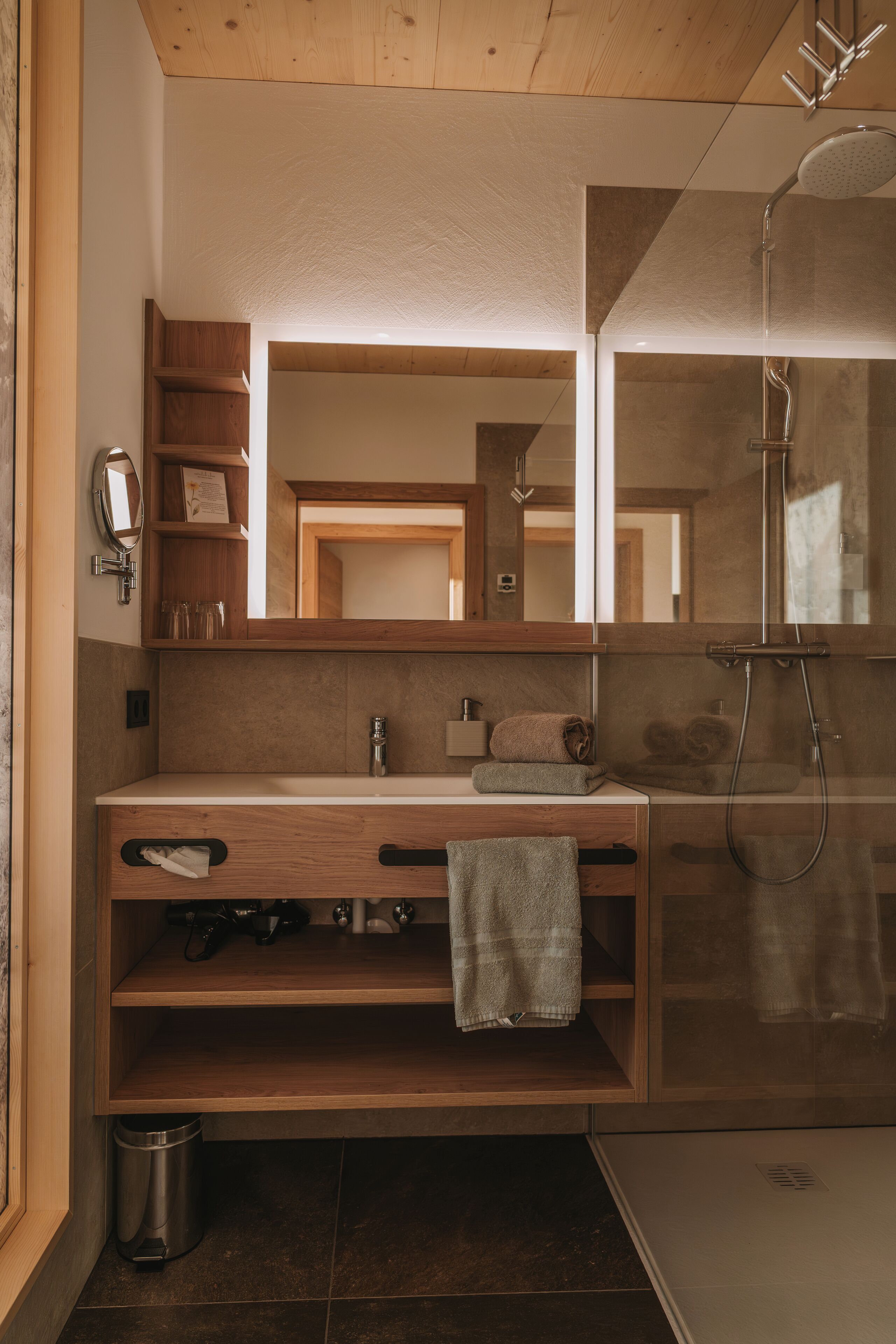 A modern bathroom with a wooden vanity, a large mirror, a shower, and a trash can.