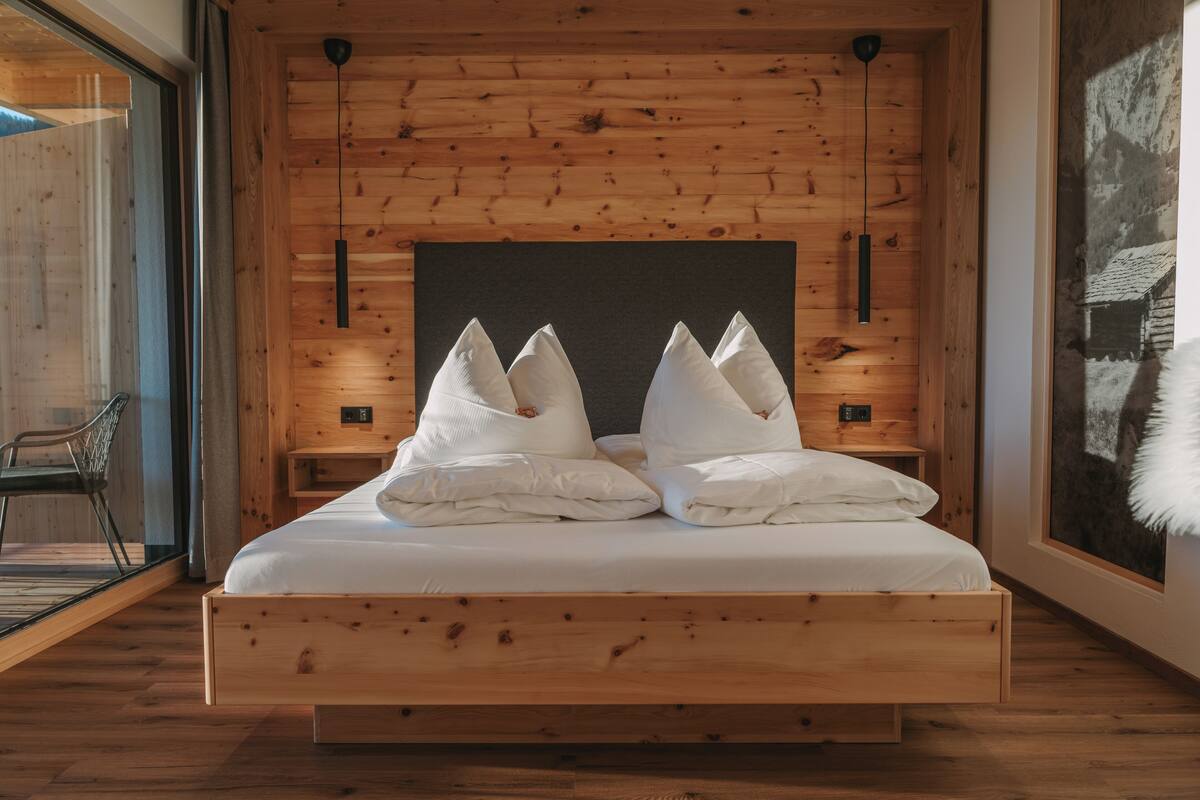Une chambre à coucher avec un cadre en bois, comprenant un lit, des oreillers et une vue sur un bâtiment rustique, visible à travers une fenêtre.