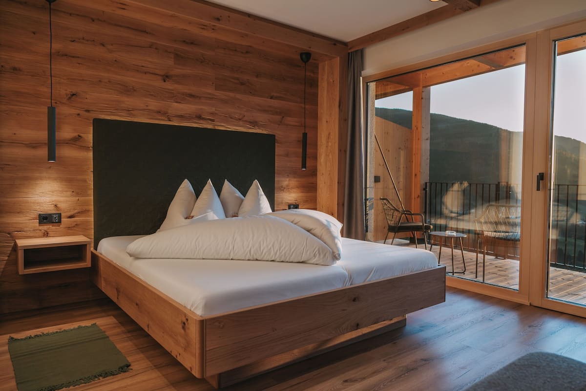 Une chambre à coucher en bois, avec un grand lit, une table de chevet, un tapis vert et une vue sur l’extérieur grâce à une porte vitrée.