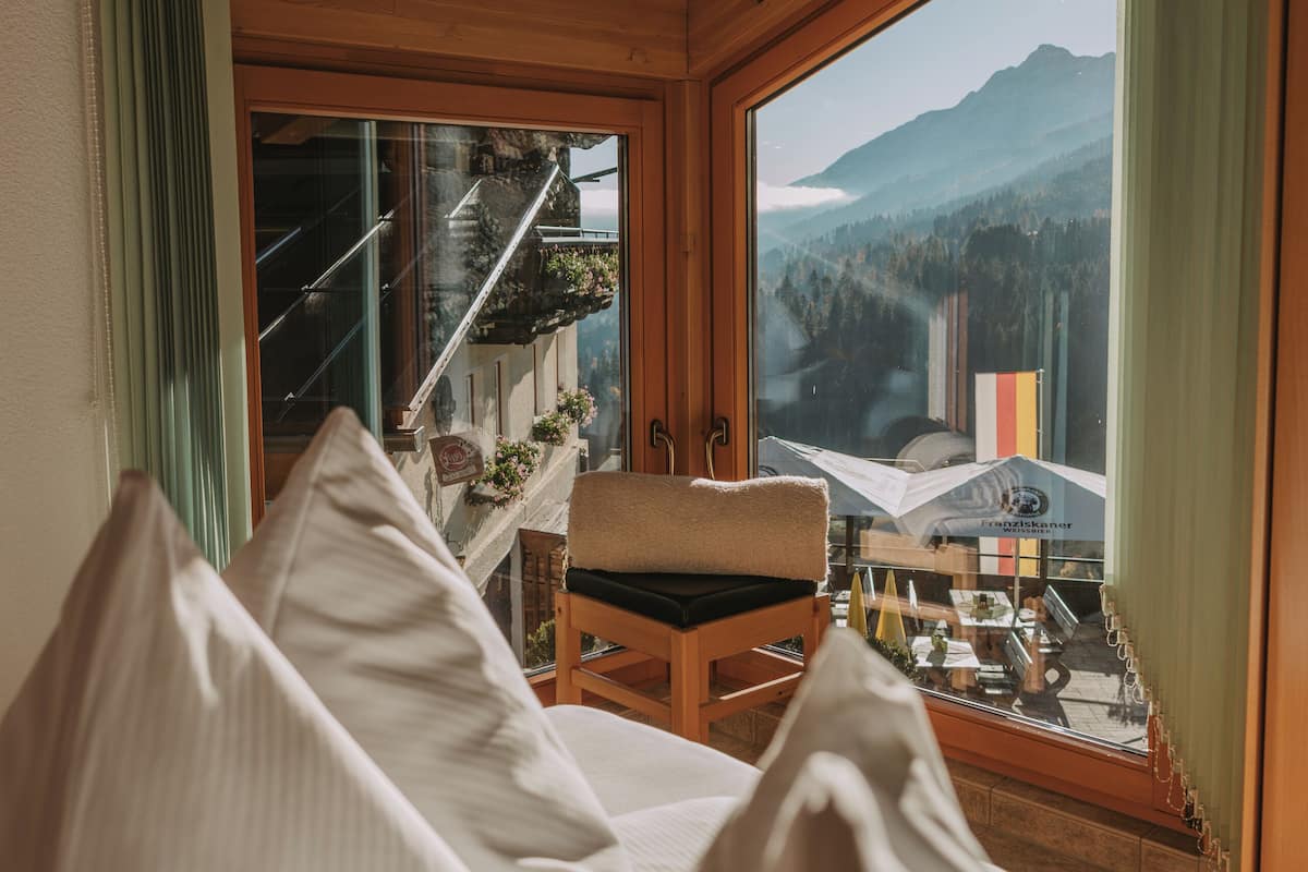 Une pièce chaleureuse avec un plafond en bois, un lit recouvert de draps blancs, une chaise et une grande fenêtre donnant sur les montagnes.