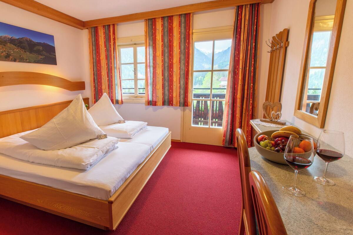 Une chambre d’hôtel avec un lit en bois, un coin repas avec des fruits et des verres à vin, et une vue sur la montagne par la fenêtre.