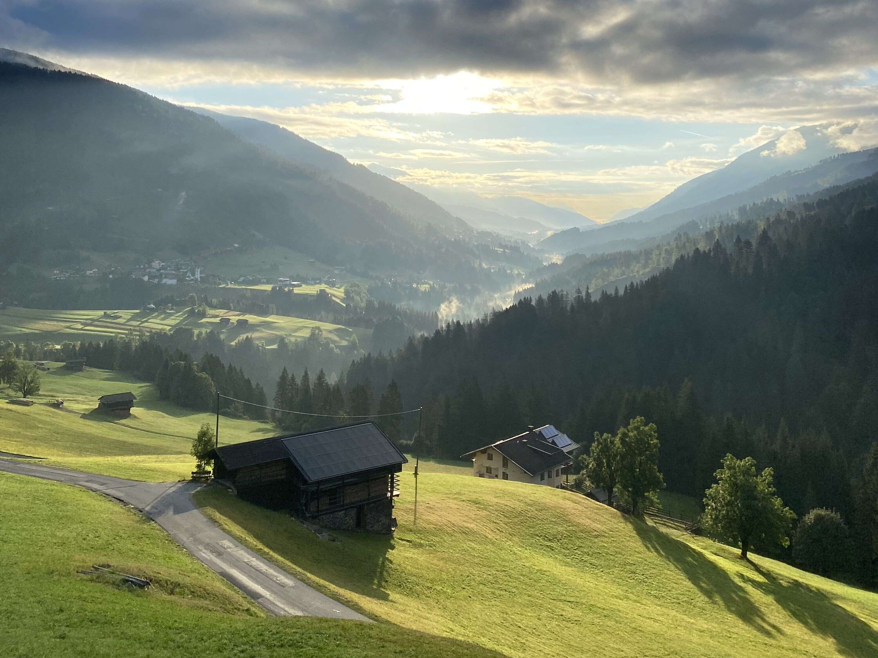 Paisaje rural con una carretera, una pequeña casa de madera y un edificio más grande con techo inclinado.