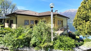 A house with a balcony overlooking a lake, surrounded by lush greenery and mountains in the background. - Apartment by Lake Maggiore with Mountain Views (Pino sulla Sponda del Lago Maggiore)