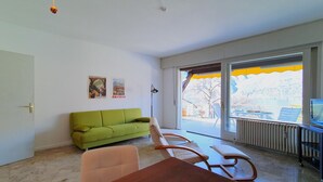 A modern living room with a green sofa, a wooden dining table, and a large window with a view of the outdoors. - Apartment by Lake Maggiore with Mountain Views (Pino sulla Sponda del Lago Maggiore)