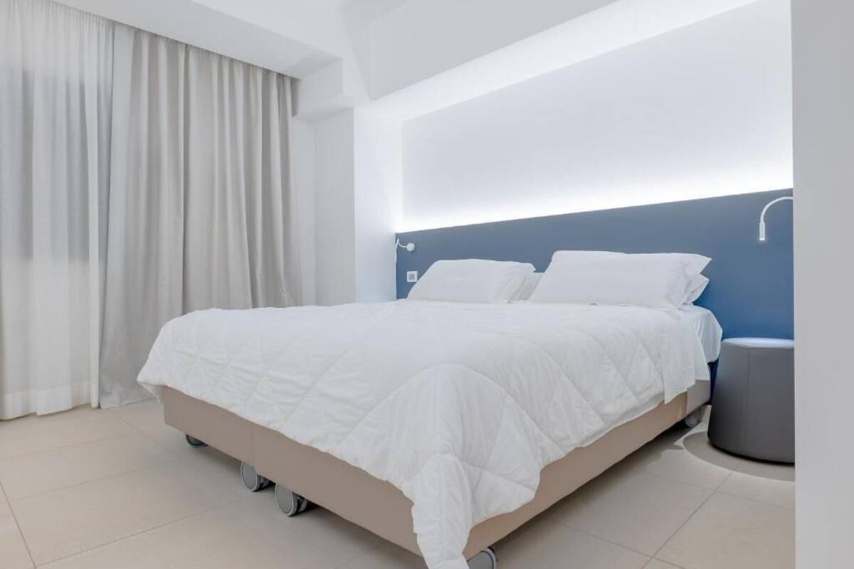 Una habitación de hotel moderna con una cama grande, cabecera con iluminación integrada, mesita de noche y ventana cubierta con cortinas.