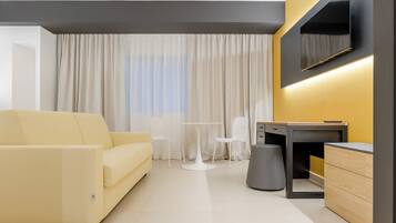 Ein modernes Hotelzimmer mit einer Couch, einem kleinen runden Tisch, zwei Stühlen, einem Schreibtisch mit Stuhl, einem an der Wand montierten Fernseher und einem großen Fenster mit Vorhängen.