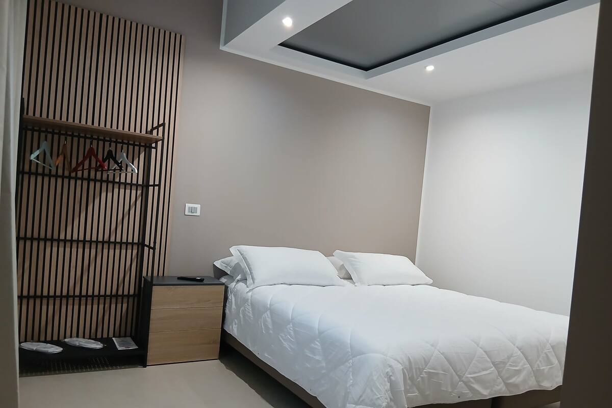 Un dormitorio con cama, mesita de noche, armario de madera y perchero con ropa.
