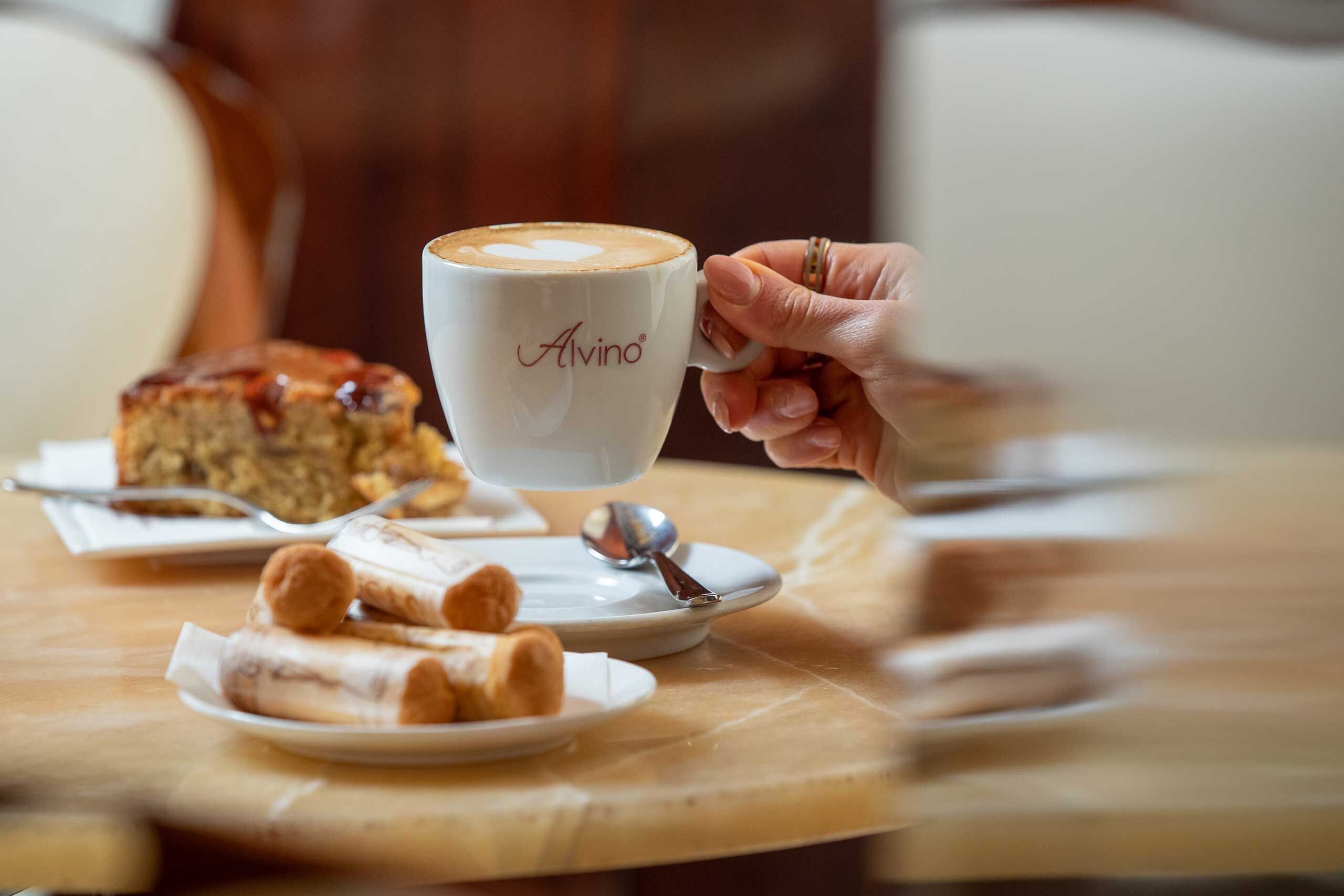 A hand holding a cup of coffee with a logo, a plate with pastries, and a cake on a table.