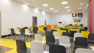Une salle de conférence avec des rangées de chaises et de tables, un tableau blanc et un logo sur le mur.