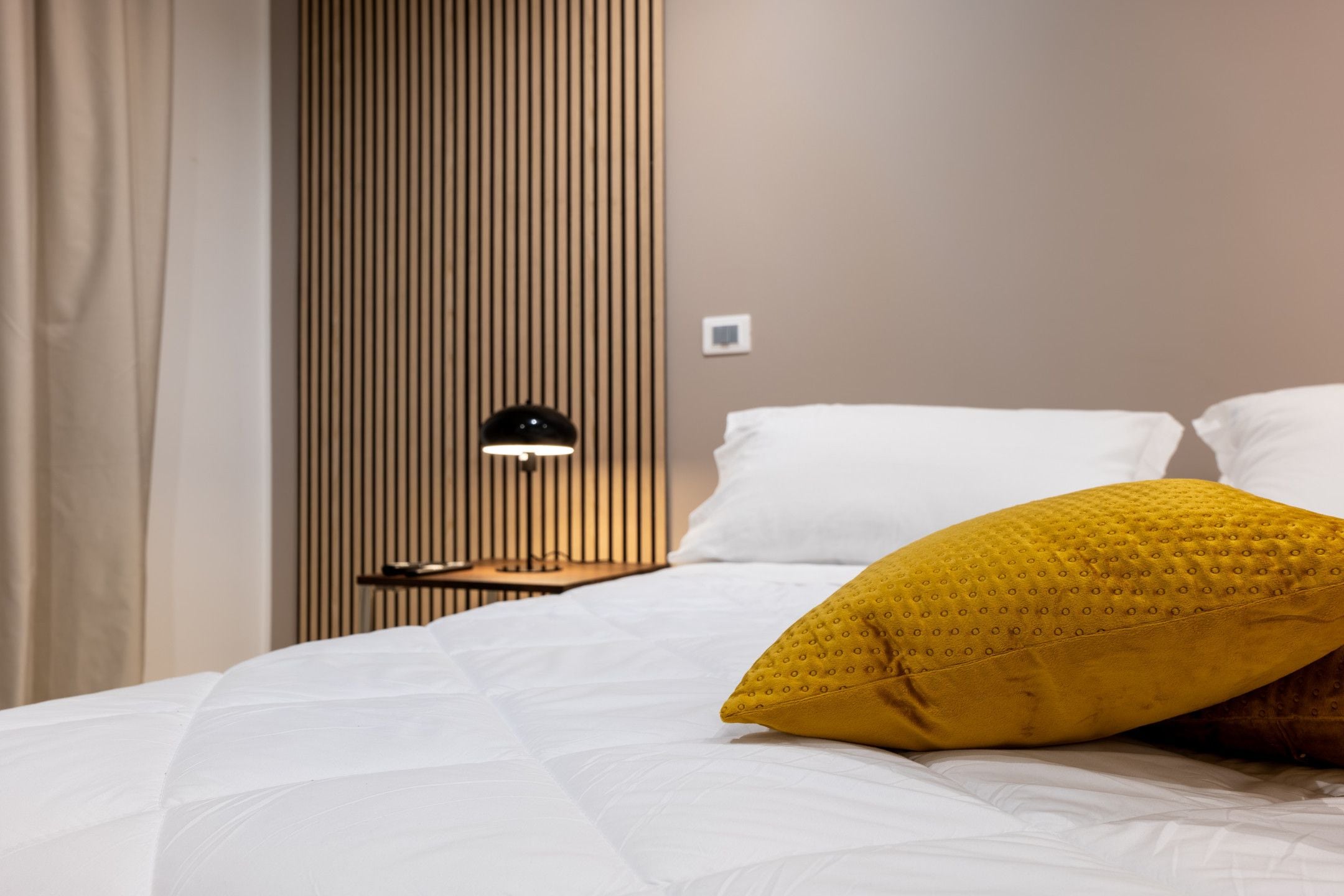 A neatly made bed with white bedding and a single yellow pillow, a bedside table with a lamp, and a vertical wooden panel wall.