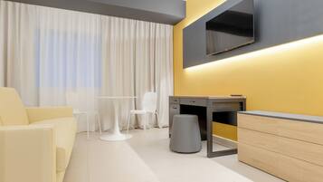 Una habitación de hotel moderna con sofá, escritorio, silla, televisor montado en la pared y un amplio ventanal con cortinas.
