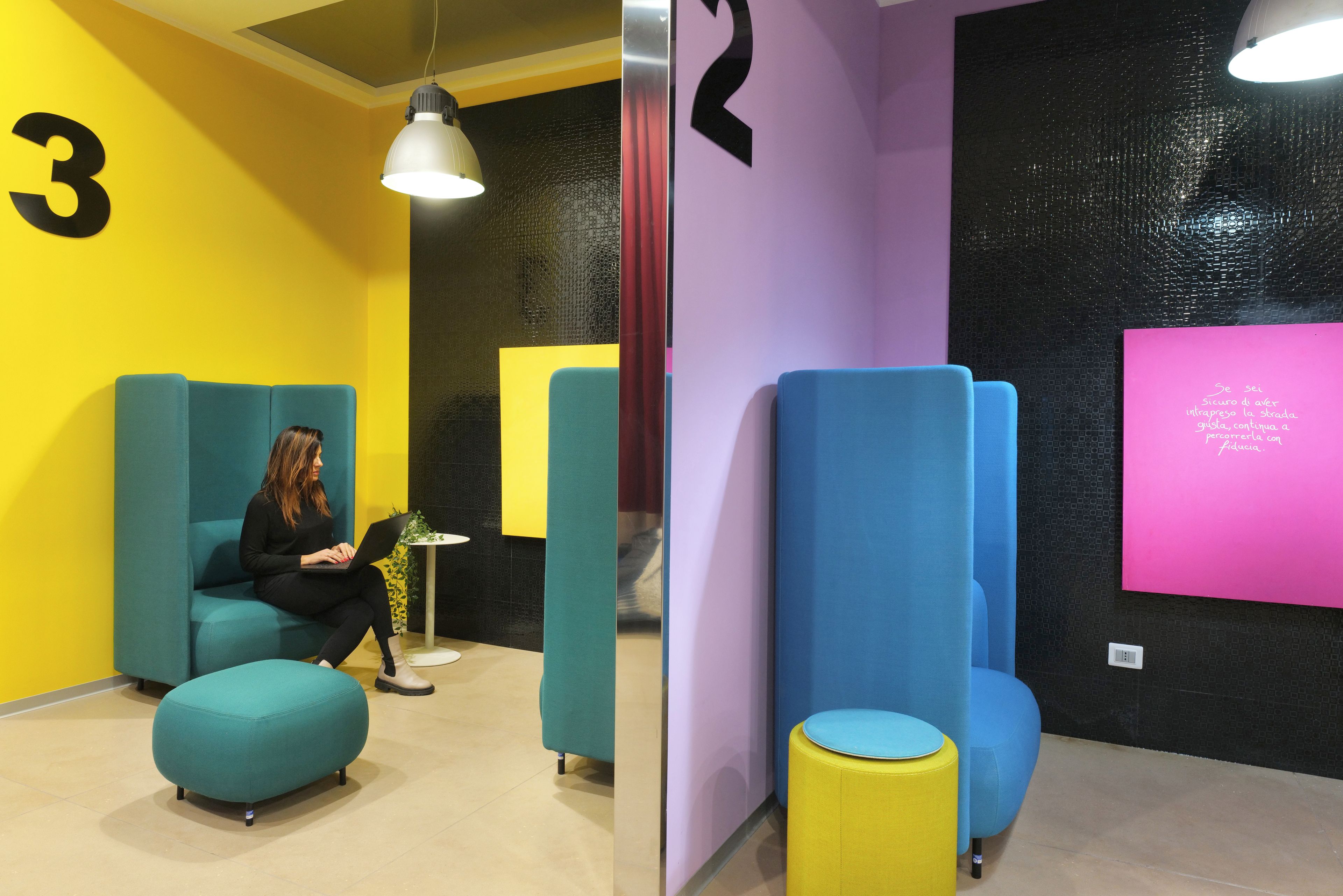 Una habitación con asientos en tonos aguamarina y amarillo, una mesa blanca y una pared rosa con texto.