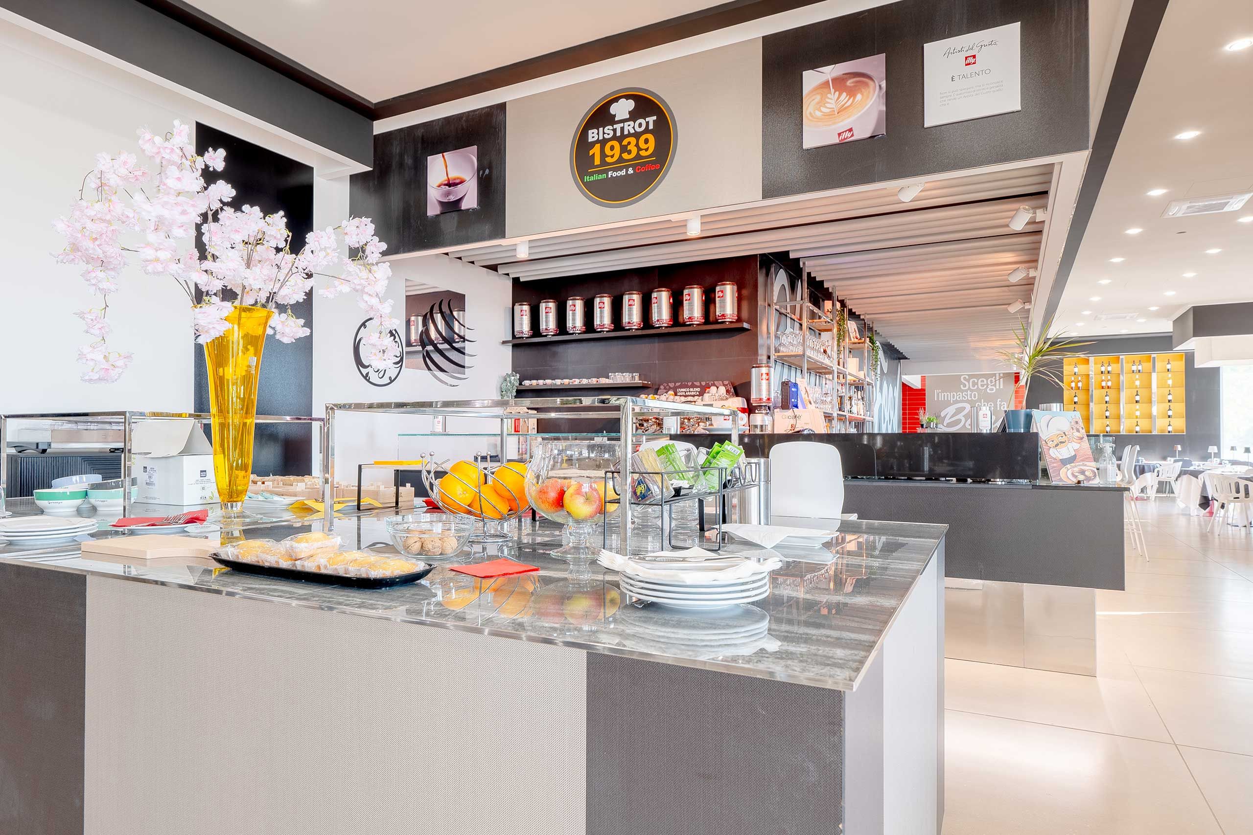 Interior de una cafetería moderna con un mostrador exhibiendo alimentos, un jarrón decorativo con flores y un letrero que indica el nombre y la especialidad de la cafetería.