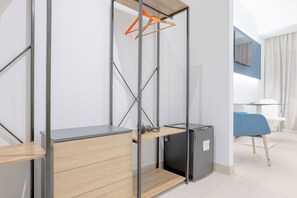 A modern hotel room with a built-in desk, a small refrigerator, a hanging clothes rack, and a seating area with a blue chair. - LHI Healthy Hotel Lecce (Lecce)