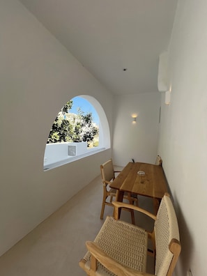 A small, cozy room with a wooden table and wicker chairs, a large arched window, and a white wall.