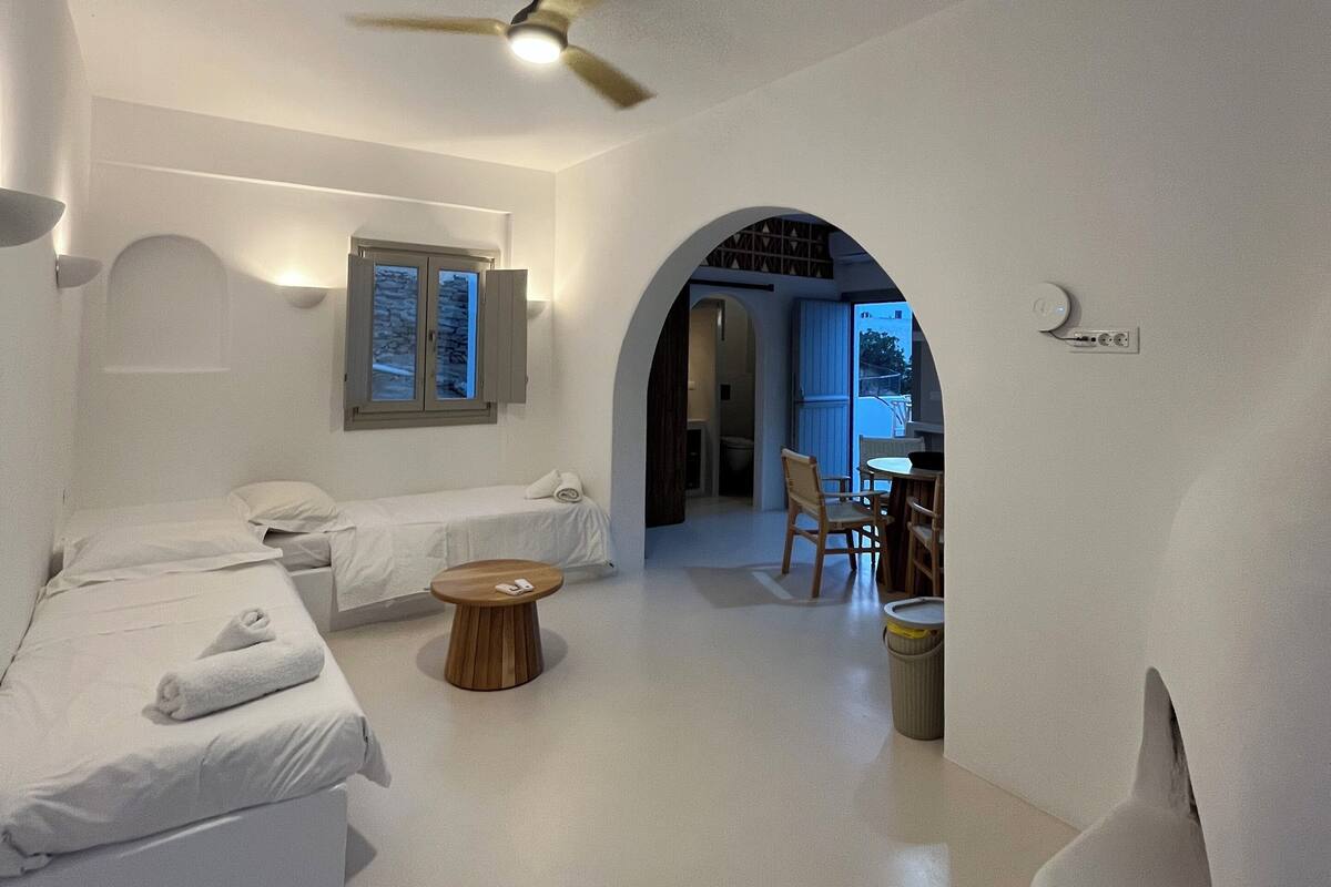 A modern, minimalist hotel room with two beds, a ceiling fan, a small round table, and a view through an arched doorway to a dining area.