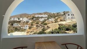 A room with a table and two chairs, a window with a view of a hillside with white buildings. - Ios Alcyon Blue Suites (Ios)