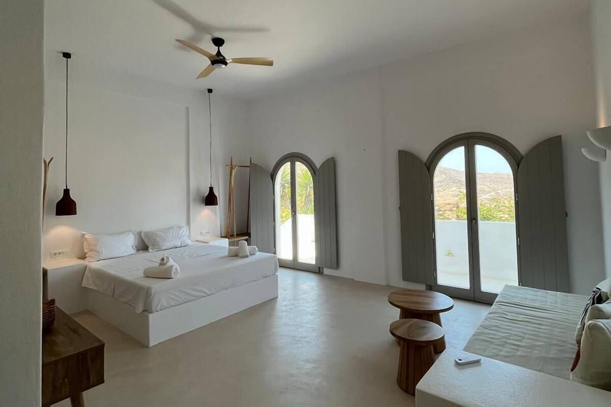 A spacious bedroom with a large bed, a ceiling fan, and a view through arched windows.