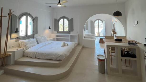 A modern, minimalist bedroom with a large bed, built-in seating area, and a dining table. - Ios Alcyon Blue Suites (Ios)