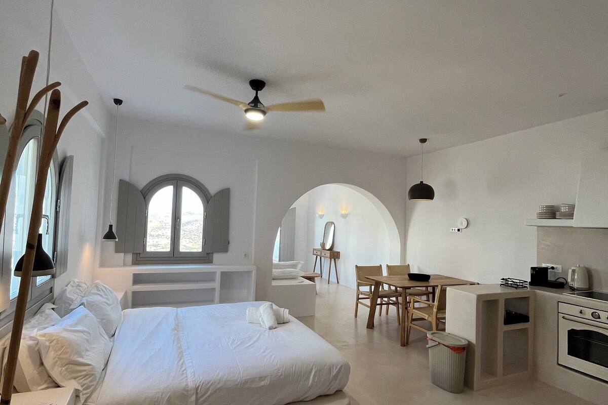 A modern, minimalist bedroom with a large bed, a small kitchen, and a dining area.